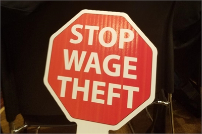 [ai] A stop sign with the message "STOP WAGE THEFT" written in bold white letters on a red background, placed on a table in a public setting.